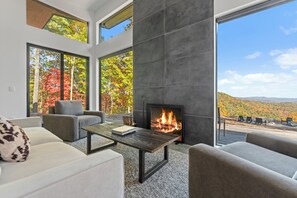 Ein modernes Wohnzimmer mit Kamin, großen Fenstern und Blick auf Herbstlaub.