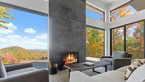 A modern living room with a fireplace, large windows, and a view of autumn foliage. - Ridge (Lenoir)