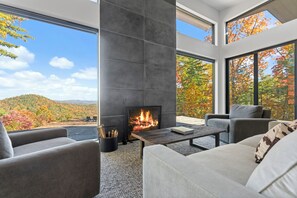 Ein modernes Wohnzimmer mit Kamin, großen Fenstern und Blick auf Herbstlaub.