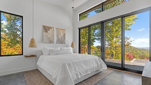 A modern bedroom with a large bed, a view of autumn foliage through floor-to-ceiling windows, and minimalist decor. - Ridge (Lenoir)
