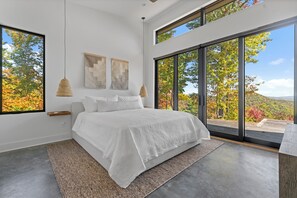 Ein modernes Schlafzimmer mit einem großen Bett, Blick auf Herbstlaub durch raumhohe Fenster und schlichter Einrichtung.