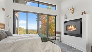 A modern bedroom with a large bed, a fireplace, and a scenic view through floor-to-ceiling windows. - Ridge (Lenoir)