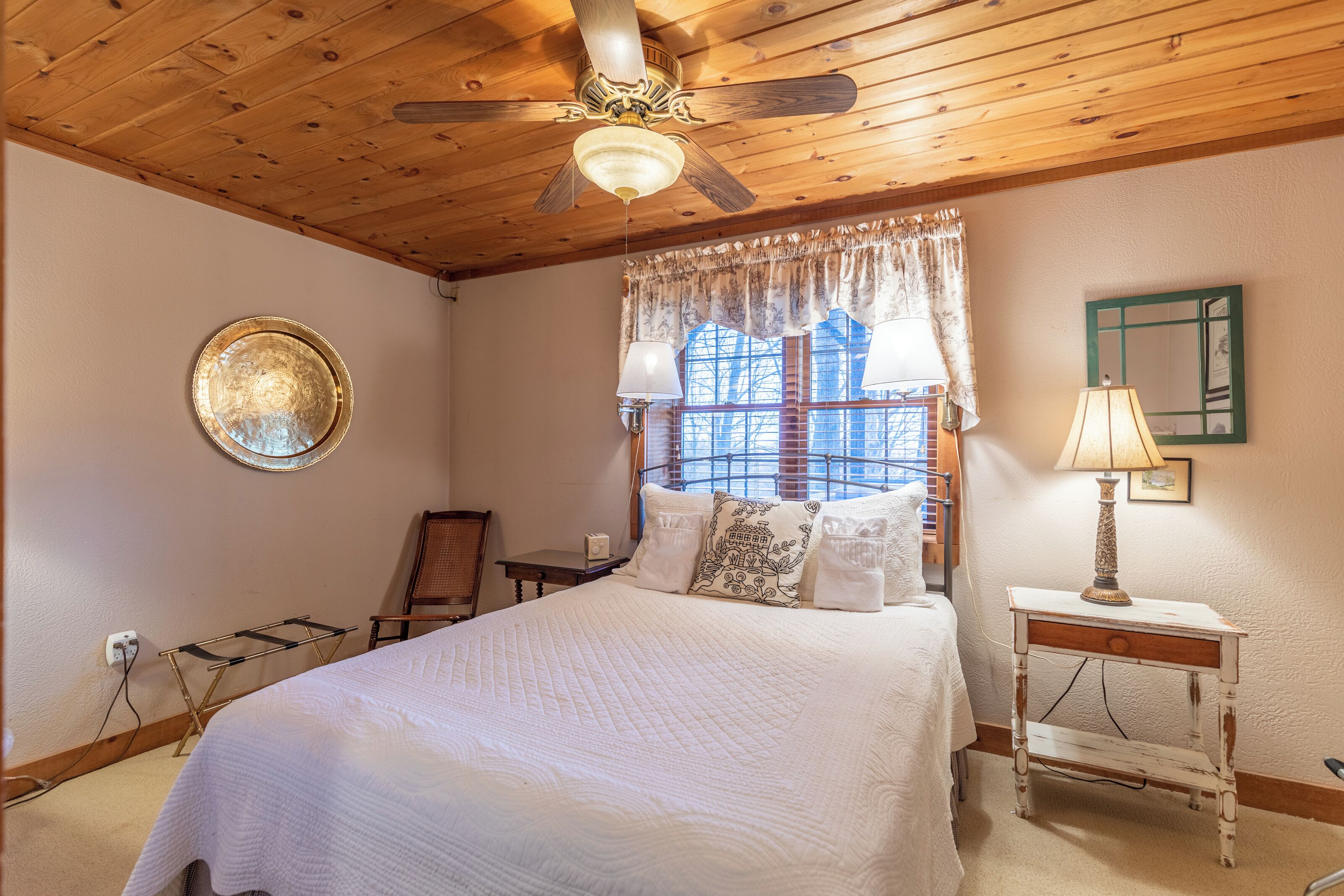 A bedroom with a wooden ceiling, a bed with white bedding, a ceiling fan, a window with lace curtains, and a bedside table with a lamp.