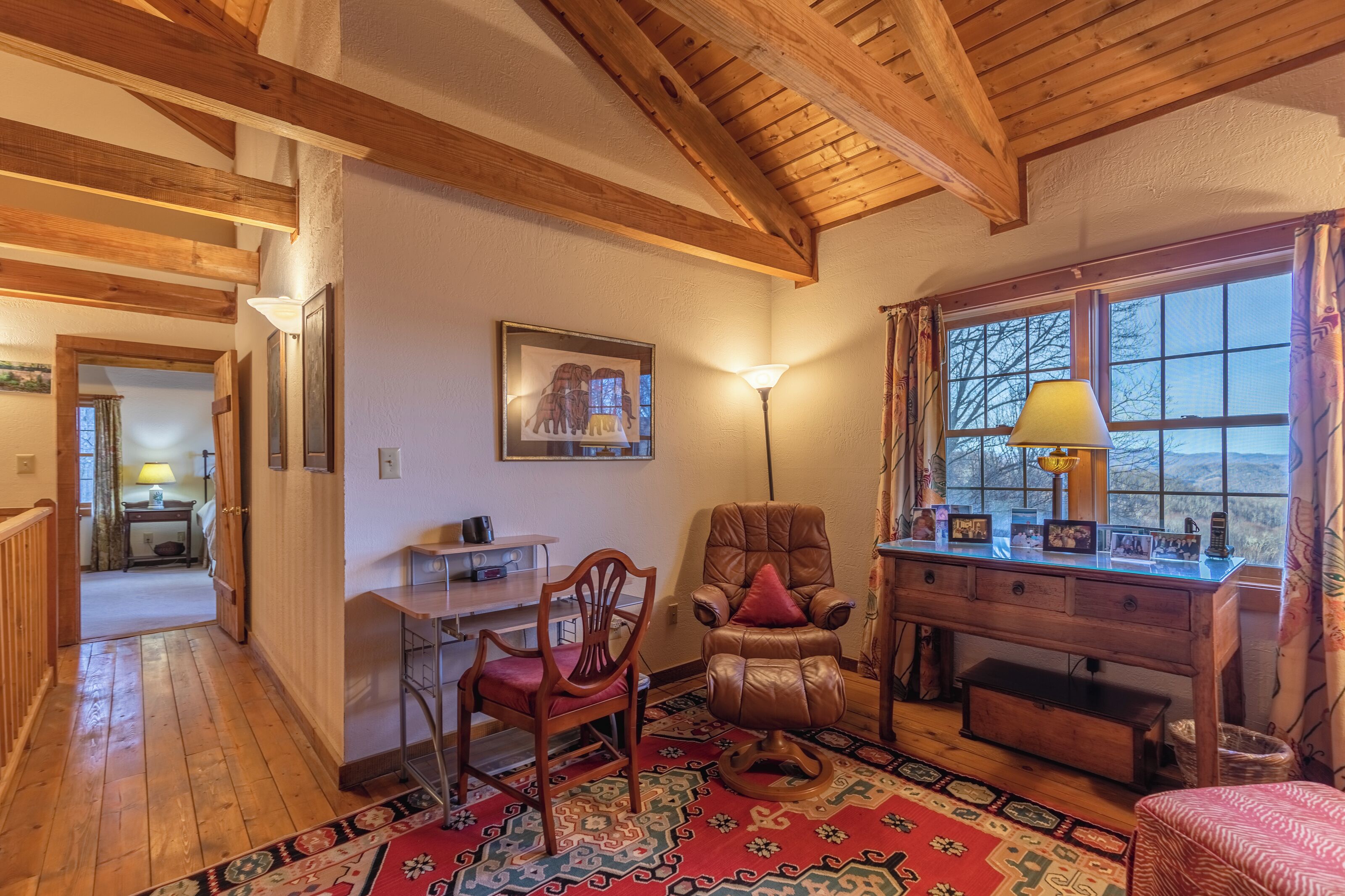 Una habitación acogedora con vigas de madera, un escritorio con lámpara, una silla y vistas por la ventana.