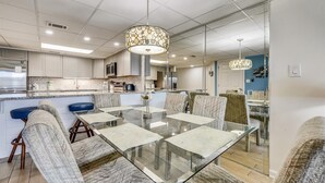 Una cocina moderna con una isla central, un comedor con mesa de vidrio y sillas acolchadas, y una sala de estar con una pared azul de acento.
