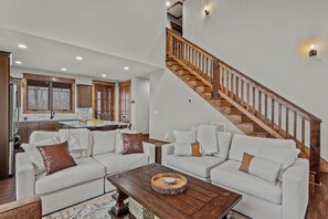 A spacious living area with a wooden staircase, white sofas, and a kitchen in the background. - Cornerstone Cabin at Eagles Nest (Banner Elk)
