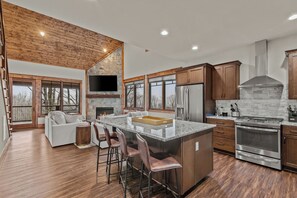 A modern kitchen with a stone fireplace, stainless steel appliances, and a large island with bar stools. - Cornerstone Cabin at Eagles Nest (Banner Elk)