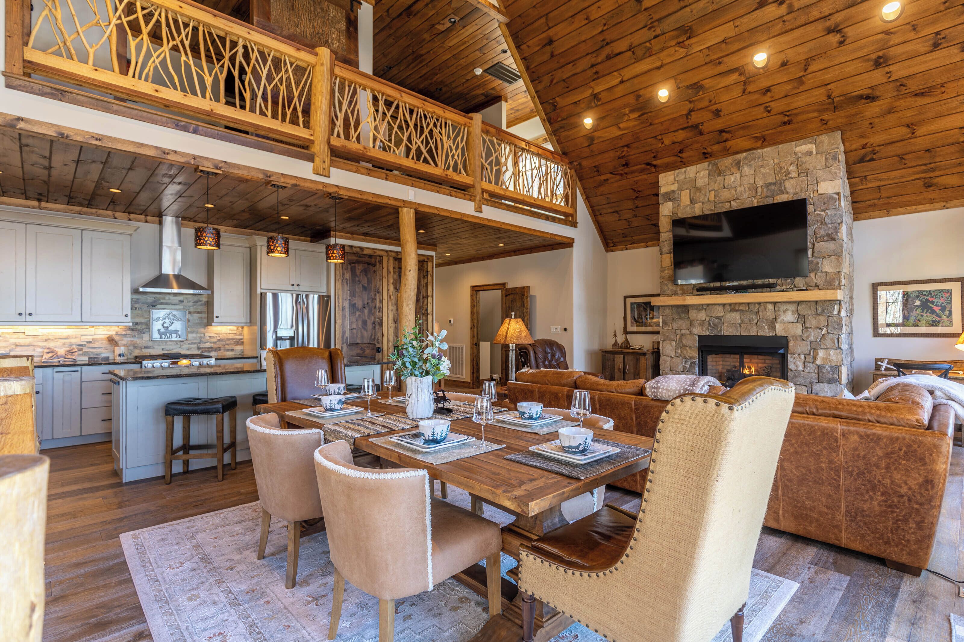 A spacious living area with a high ceiling, wooden beams, a stone fireplace, a television mounted on the wall, and a dining area with a table set for four.
