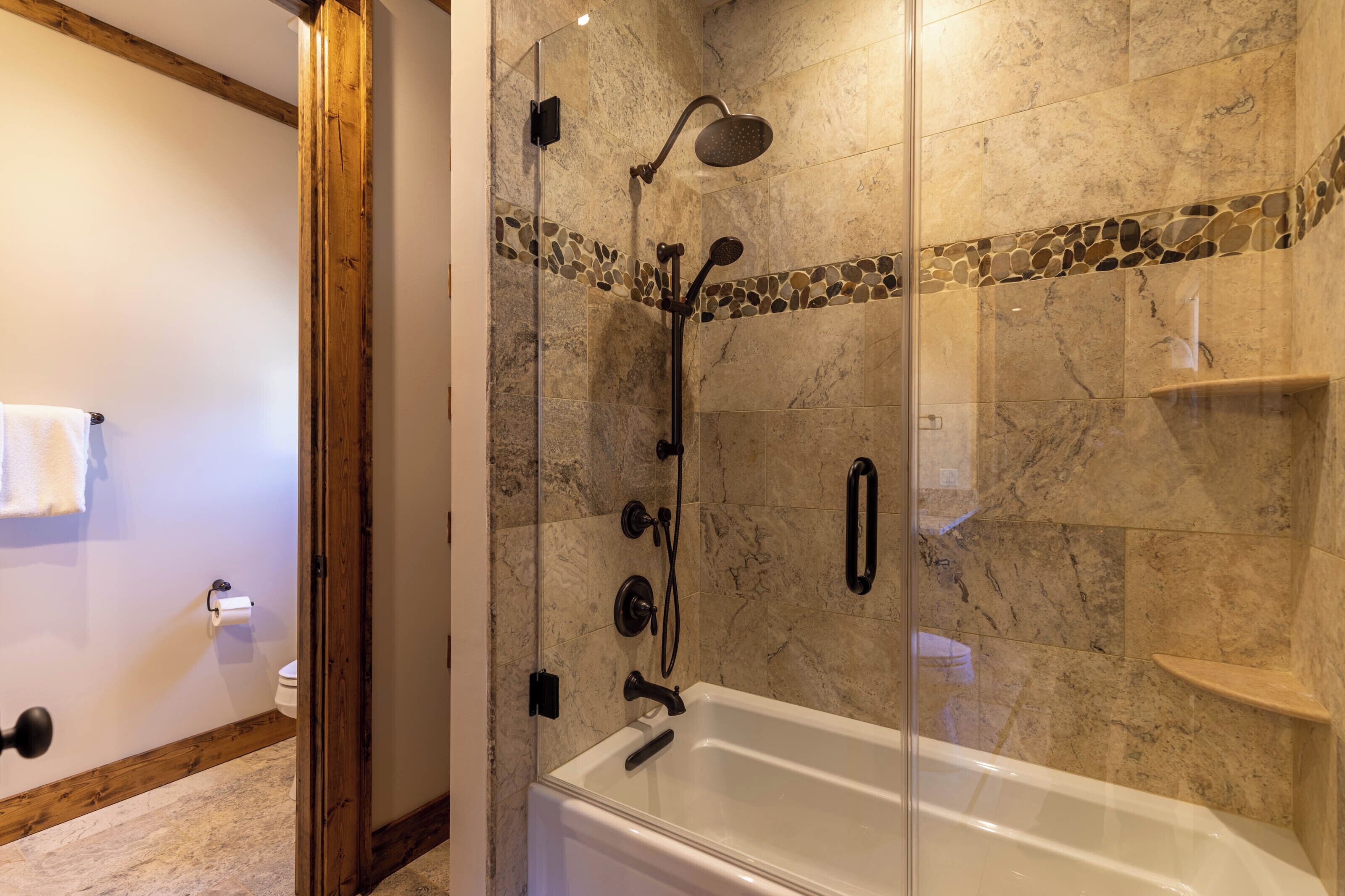 A modern bathroom with a glass-enclosed shower, a white bathtub, and a stone mosaic border.