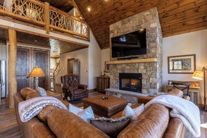 A spacious living room with a stone fireplace, leather sofas, a television mounted above the fireplace, and a wooden ceiling.