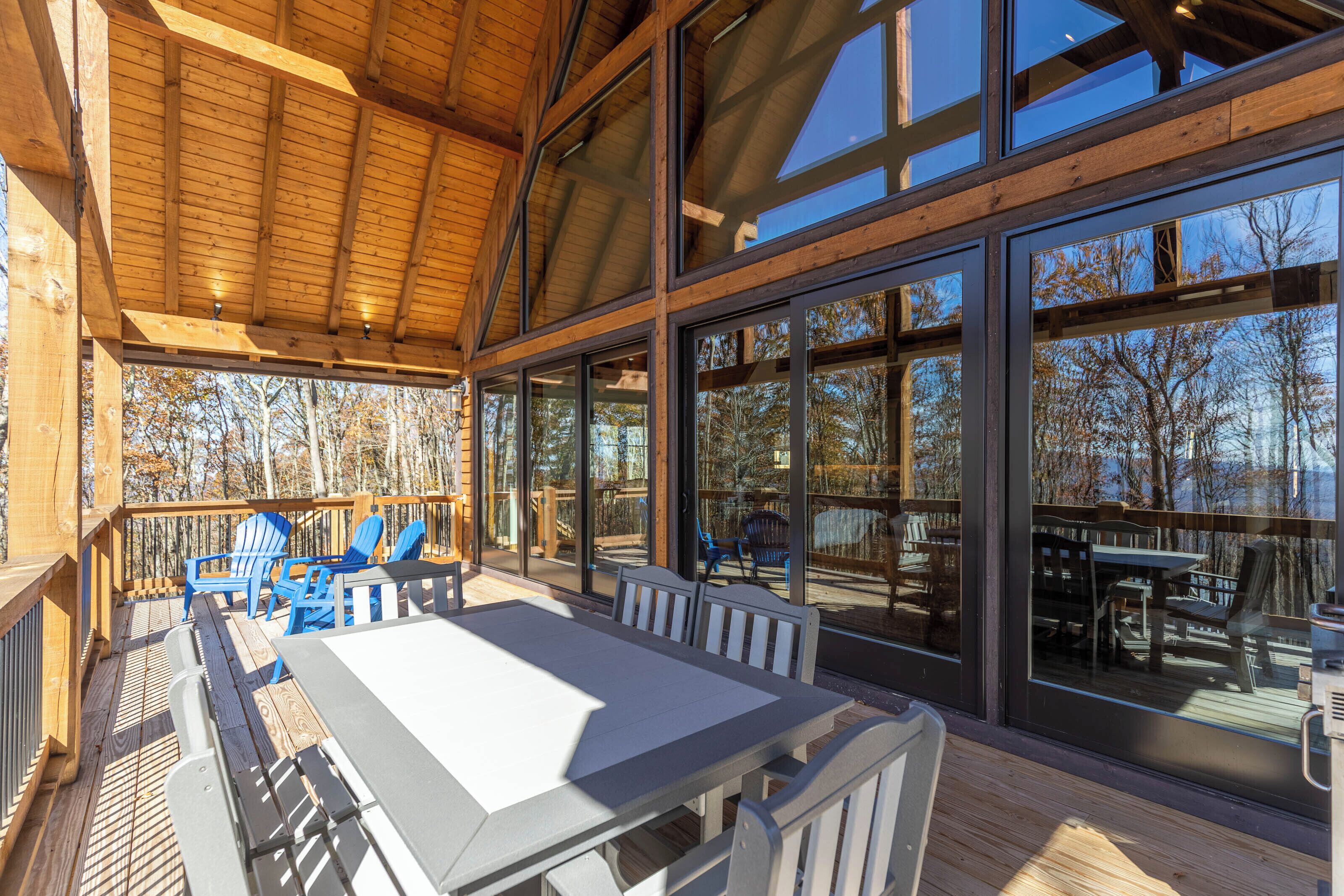 A covered outdoor deck with a table and chairs, large windows, and a view of trees.