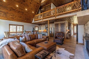 A spacious living room with a high ceiling and wooden beams, a cozy fireplace, and a dining area with a kitchen in the background.
