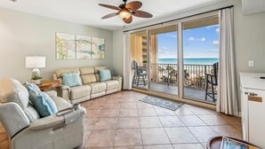 A spacious living room with a beige sofa, a gray armchair, and a ceiling fan. A large sliding glass door leads to a balcony with a view of the beach. - Shores of Panama 521 (Panama City Beach)