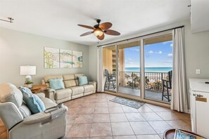 Ein geräumiges Wohnzimmer mit einem beigen Sofa, einem grauen Sessel und einem Deckenventilator. Eine große Glasschiebetür führt zu einem Balkon mit Blick auf den Strand.