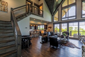 A spacious living area with a high ceiling, a staircase, a kitchen, and large windows offering a view of the outdoors. - Just Bluffin' at Eagles Nest (Banner Elk)