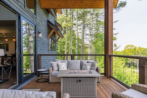 Eine überdachte Terrasse mit Feuerstelle, Sitzgelegenheiten und Blick auf einen Wald.