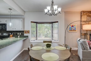A dining area with a round table, chairs, a chandelier, a stone fireplace, and a window with blinds.
