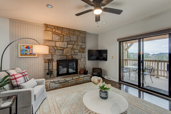 A cozy living room with a stone fireplace, a flat-screen TV, a comfortable sofa, and a dining area with outdoor seating. - Chetola Robin 1 (Blowing Rock)