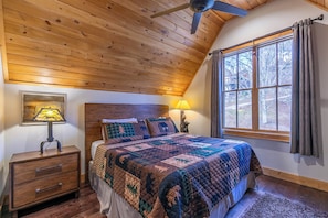 Ein Schlafzimmer mit Holzboden, einem großen Fenster mit Vorhängen, einem Bett mit Quilzdecke, einem Nachttisch mit Lampe und einem Wandbild.
