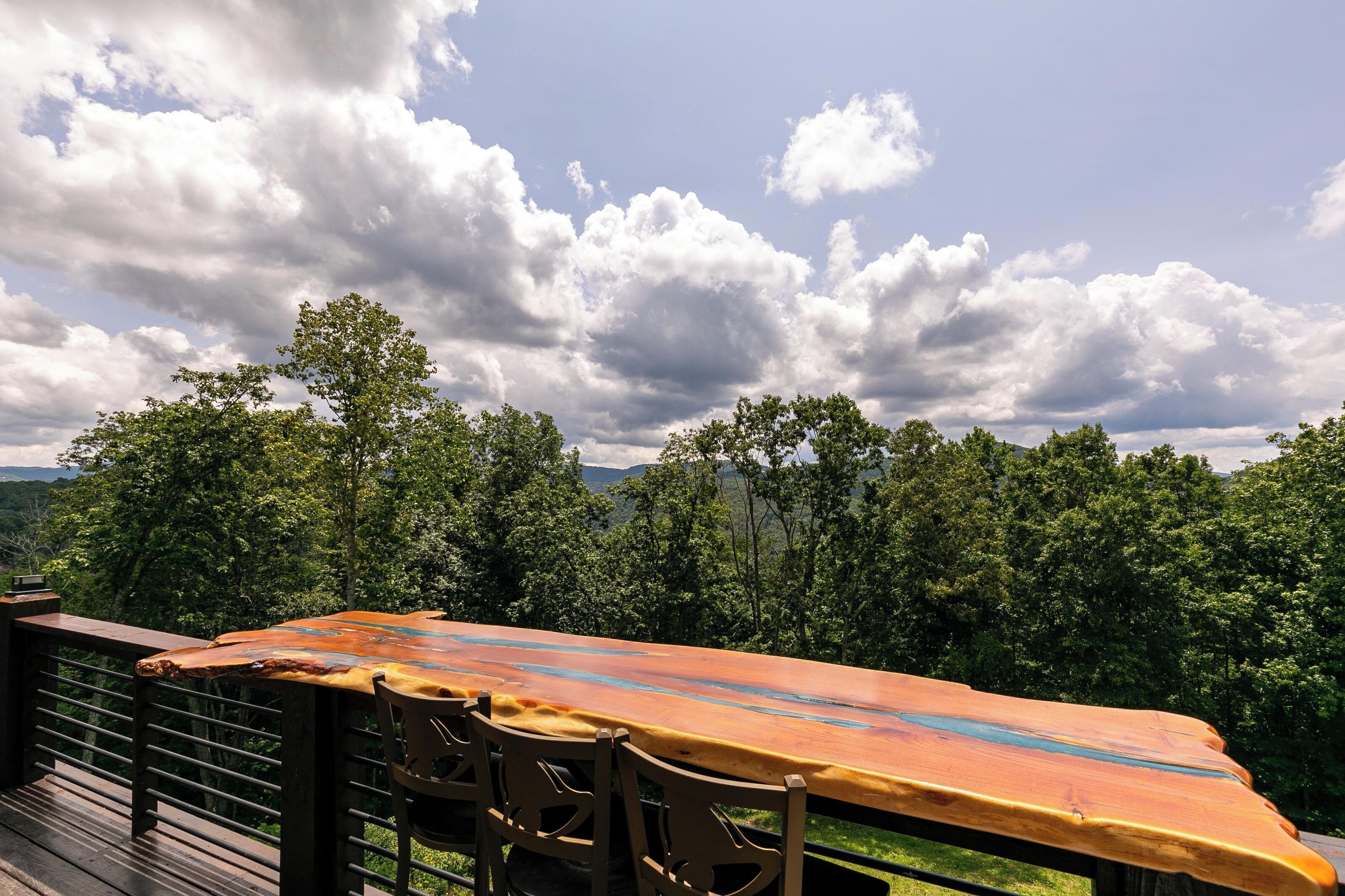 Une terrasse en bois avec une table et des chaises, offrant une vue sur un paysage boisé et un ciel nuageux.