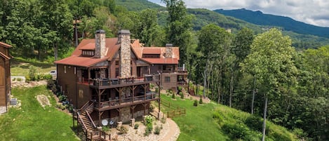 A large, rustic cabin with a stone chimney, multiple decks, and a stone fireplace surrounded by a well-maintained garden.
