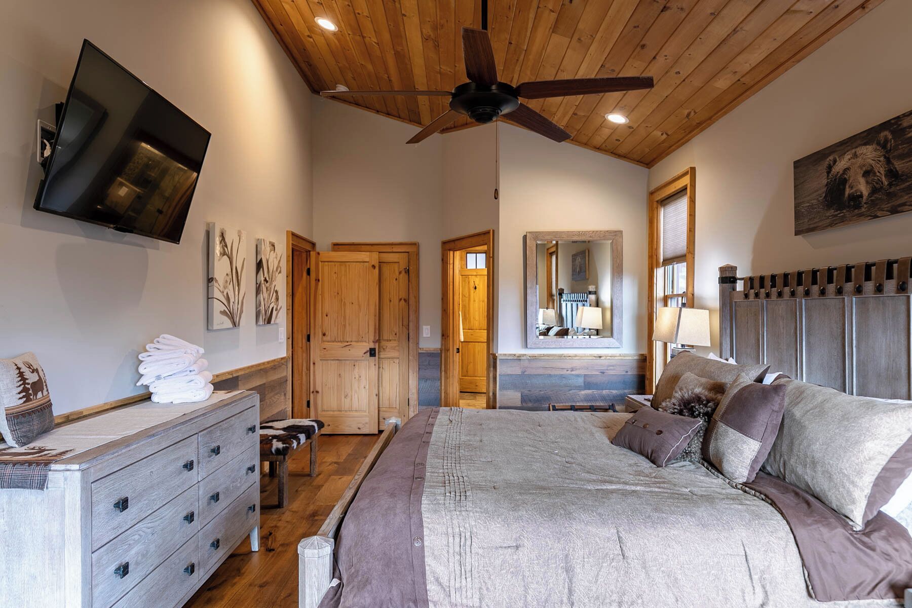 A bedroom with a wooden ceiling, a bed with multiple pillows, a dresser, and a television mounted on the wall.