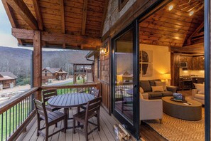 Um deck de madeira com mesa, cadeiras, um sofá e uma cozinha visível através de uma porta de vidro.
