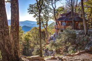 A rustic cabin with a stone foundation and wooden beams, surrounded by dense forest and rocky terrain, overlooking a mountainous landscape. - Peak A View at Eagles Nest (Banner Elk)