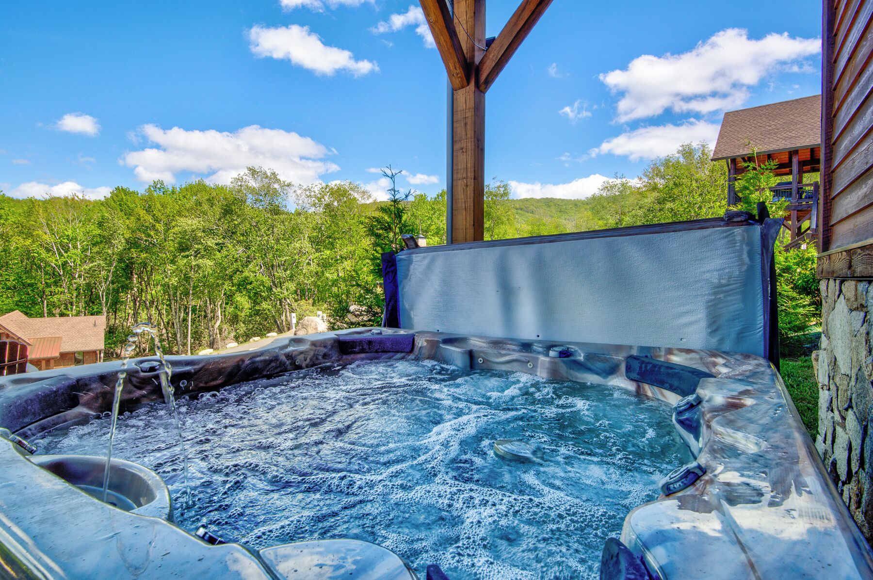 A hot tub with a wooden deck and a stone wall, surrounded by trees and a clear sky.