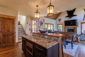 A spacious kitchen with granite countertops, wooden cabinets, and a large island. There is a fireplace, a television mounted above it, and a stone fireplace surround.