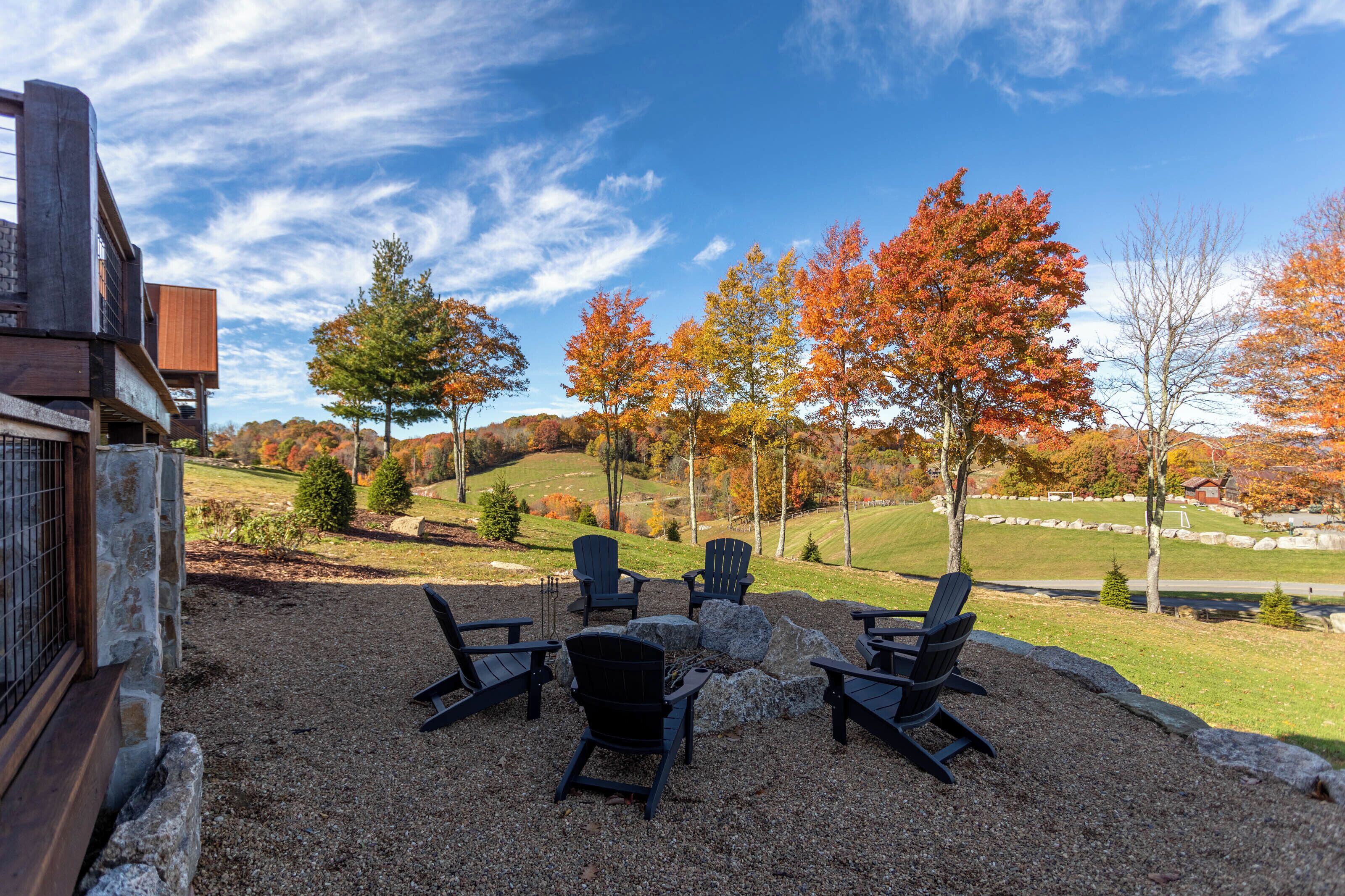 An outdoor seating area with Adirondack chairs and a fire pit, surrounded by autumn foliage and a view of rolling hills.