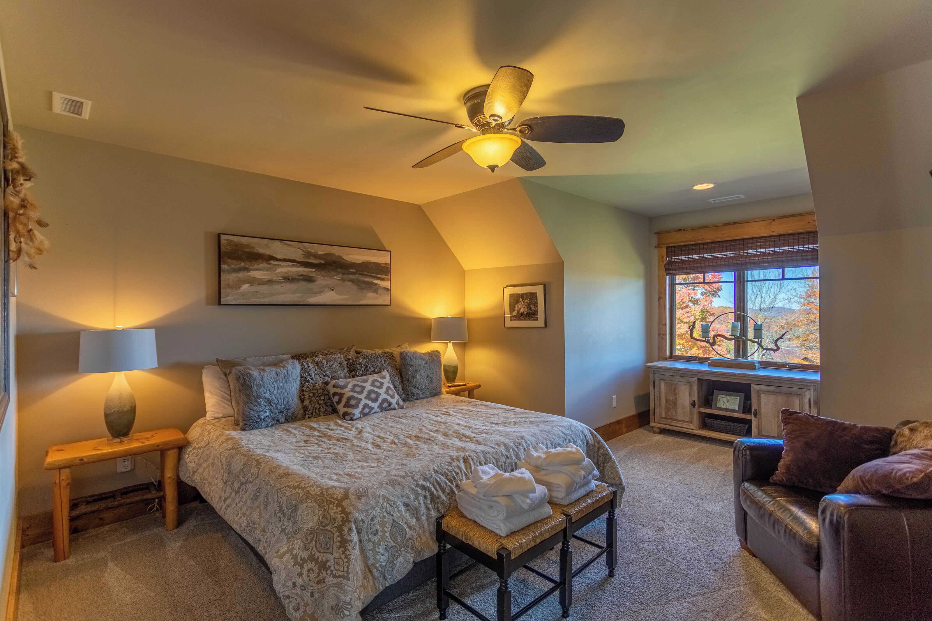 A bedroom with a large bed, two bedside lamps, a ceiling fan, a window with a view, and a bench with towels.