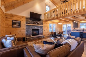 A cozy log cabin interior with a stone fireplace, leather sofas, a dining area, and a kitchen with a large window.