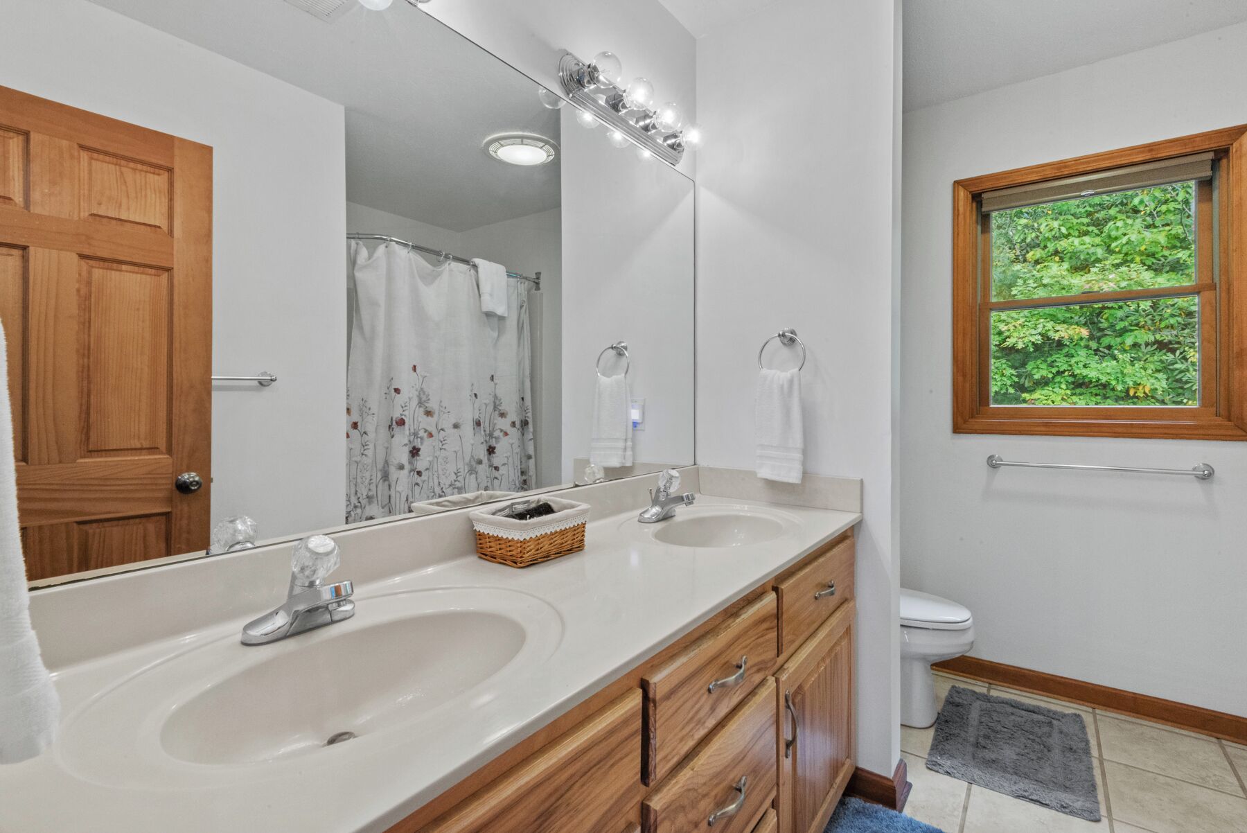 A bathroom with two sinks, a large mirror, a shower curtain, and a window with a view of greenery.