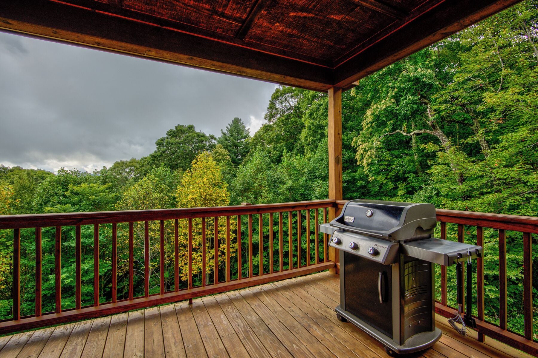 A covered outdoor area with a barbecue grill, wooden deck, and a view of dense forest.