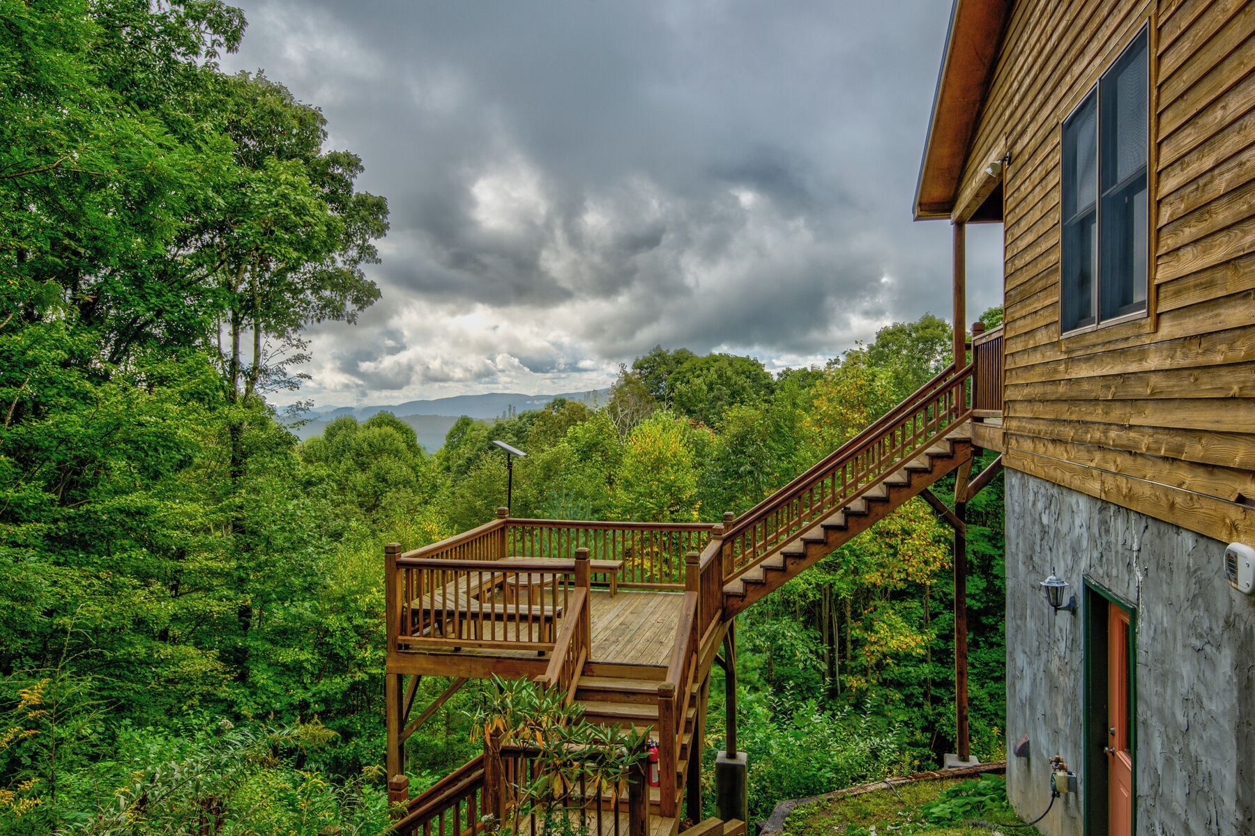 A wooden cabin with a wooden staircase leading to a deck, surrounded by dense forest and a cloudy sky.