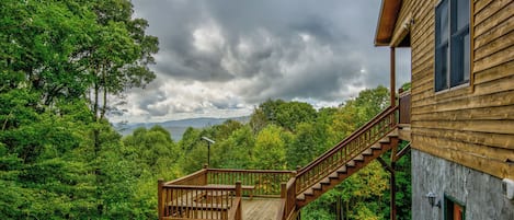 A wooden cabin with a wooden staircase leading to a deck, surrounded by dense forest and a cloudy sky.