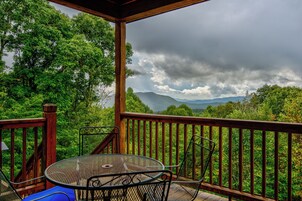 Un espacio al aire libre techado con una mesa y sillas de metal, rodeado por una barandilla de madera, con vistas a un paisaje boscoso y montañas distantes.