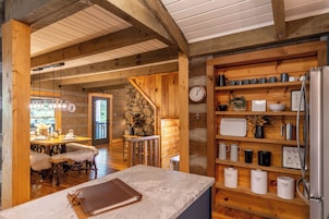 A rustic wooden interior with a stone wall, wooden shelves with kitchenware, a clock, and a dining area with a table and chairs.