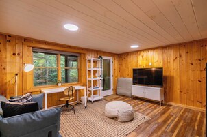 Habitación con paneles de madera, un escritorio, una silla, un reposapiés, un televisor y una ventana con vistas a un bosque.