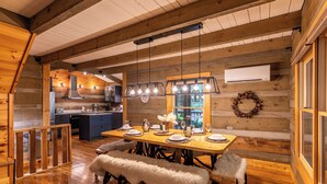 A cozy log cabin interior with a wooden dining table set for four, a kitchen area, and a large window offering a view of the outdoors. - Blackberry Creek Cabin (Lenoir)