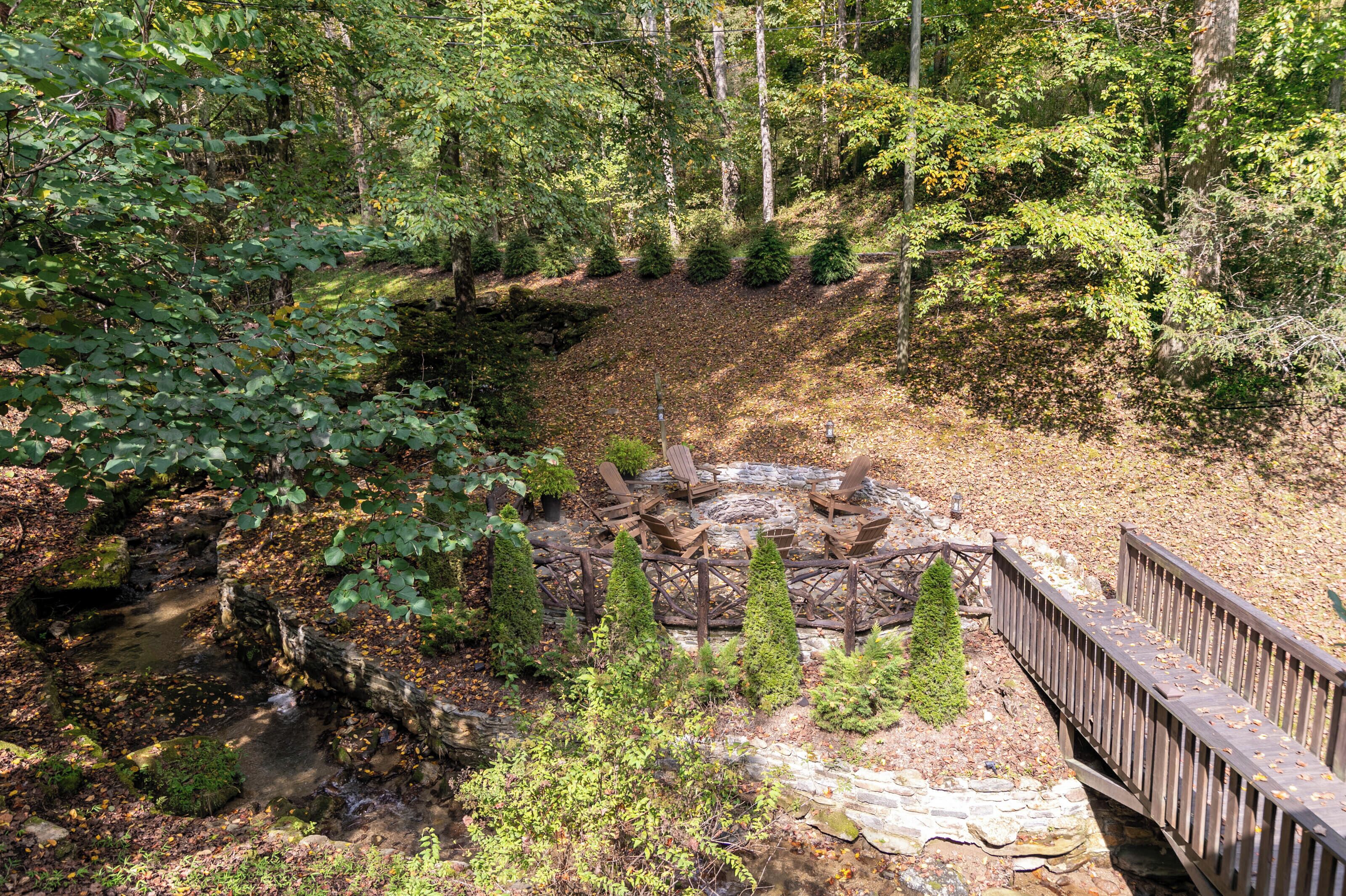 A forested area with a wooden bridge, a stone fire pit with chairs, and a small stream.