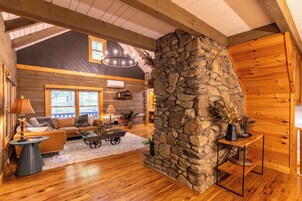 Interior de una cabaña de troncos con chimenea de piedra, vigas de madera, un sofá y una mesa de comedor.