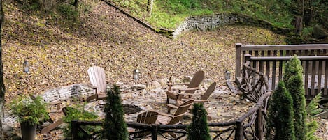En træterrasse med Adirondack-stole, omgivet af en stenvæg og et træhegn, beliggende i et skovområde med nedfaldne blade.