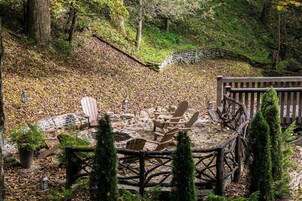 Un muelle de madera con sillas Adirondack, rodeado por un muro de piedra y una cerca de madera, ubicado en un área boscosa con hojas caídas.