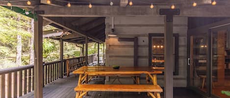 Una terraza de madera al aire libre con una mesa de picnic, luces colgantes y un vidrio que da a un edificio.