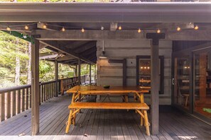 Une terrasse extérieure en bois, avec une table de pique-nique, des guirlandes lumineuses et une porte vitrée donnant accès à un bâtiment.