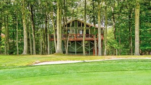 Eine zweistöckige Holzhütte mit einer Terrasse, umgeben von hohen Bäumen und einem gepflegten Rasen.