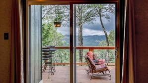 Eine Holzterrasse mit Stuhl und Tisch, eine Schiebetür aus Glas und Blick auf Bäume und Berge.