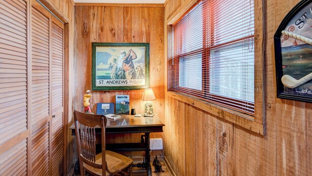 Ein Zimmer mit Holzwänden, einem Schreibtisch, einem Stuhl, einem Bilderrahmen mit einem Motiv von St. Andrews und einem Fenster mit Jalousien.